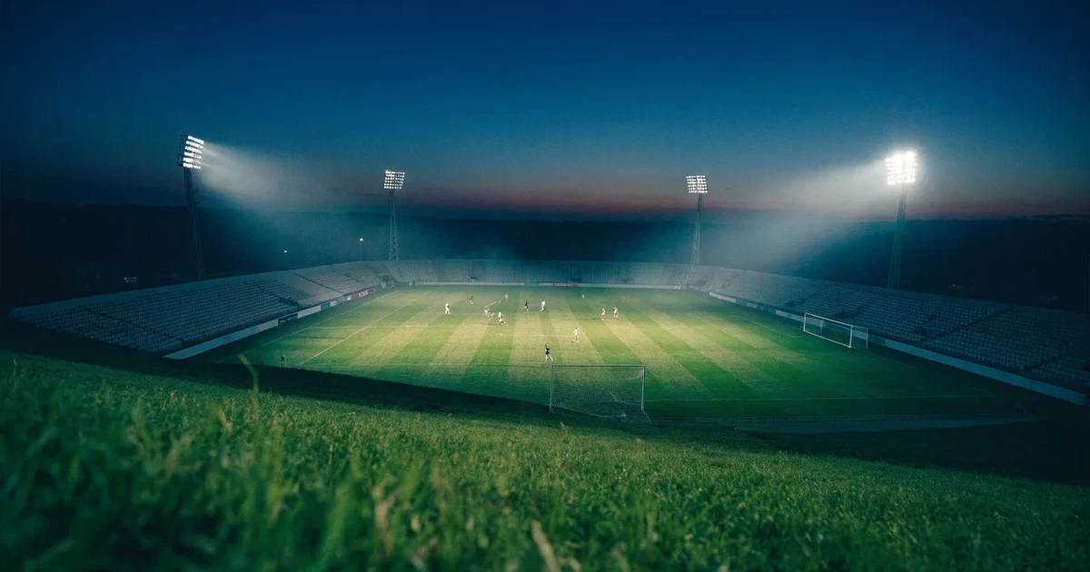 Ein Fußballstadion unter Flutlicht mit hellen Rängen, von einem Hügel aus gesehen, darüber ein dunkelblauer Abendhimmel.
